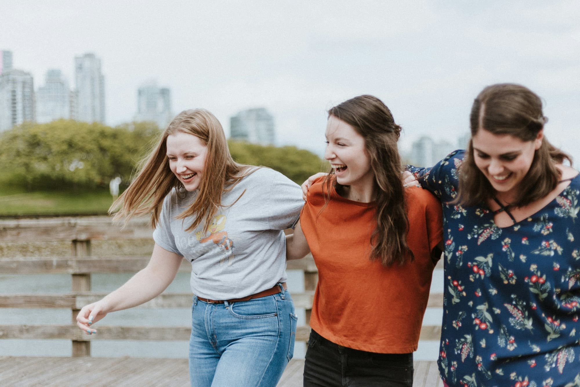 Three young women smiling and walking arm-in-arm outdoors near a waterfront with city buildings in the background